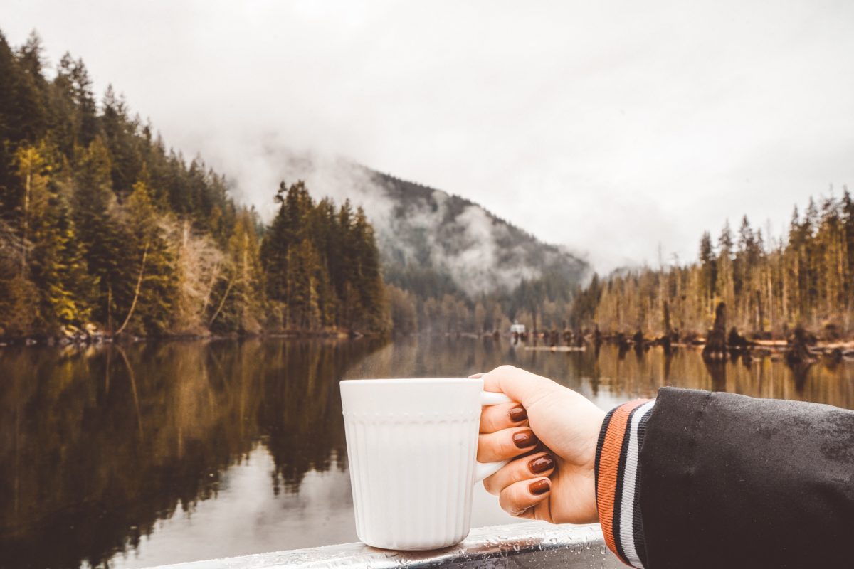 A white cup of coffee is being held in front of a lake with trees and mountains